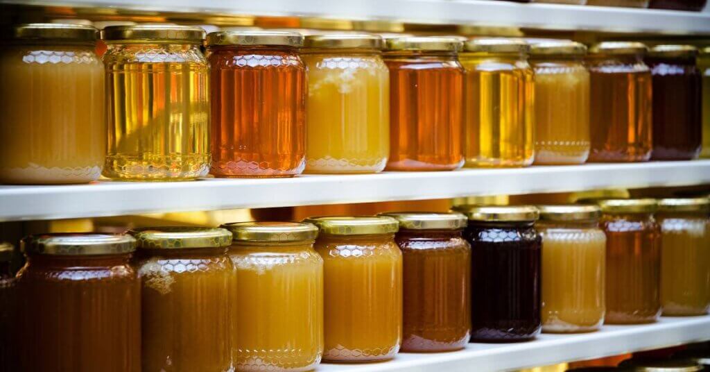 Rows of glass honey jars with metal lids on a white shelf. Each jar's honey is a different color and thickness.