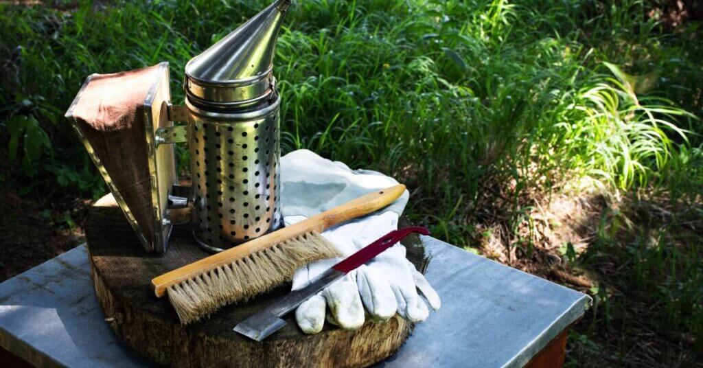 Beekeeping supplies sitting on a wooden table. There is a smoker, a bee brush, a hive tool, and gloves.