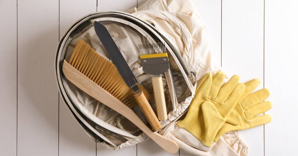 Beekeeping gear on a white paneled background. There is a folded up suit, a veil, yellow gloves, hive tools, and a bee brush.