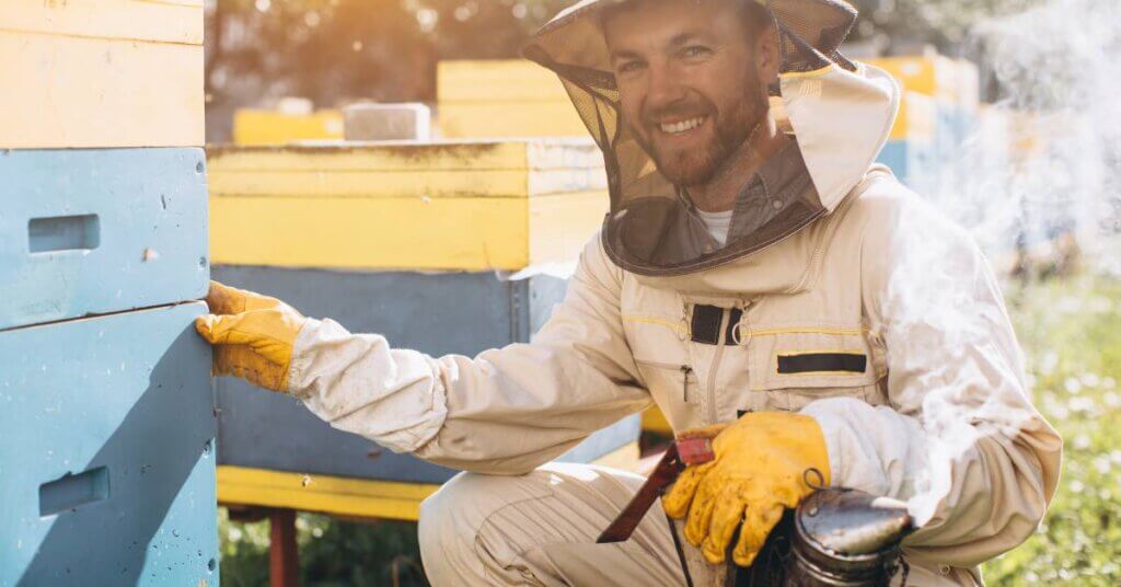 A smiling beekeeper kneeling beside a beehive. He is wearing a suit, veil, and gloves and holding a smoker.