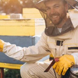 A smiling beekeeper kneeling beside a beehive. He is wearing a suit, veil, and gloves and holding a smoker.